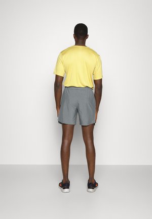 Gul kortærmet skjorte og grå shorts. Skjorten har en rund halsudskæring; shortsene er knælange med en glat overflade.