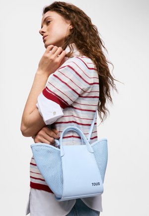 Mujer con cabello largo y rizado que lleva una camisa a rayas rojas y blancas, sosteniendo un bolso TOUS azul claro sobre su hombro.