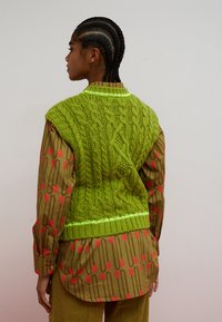 Persona con capelli intrecciati che indossa un maglione verde brillante senza maniche a trecce sopra una camicia a maniche lunghe a motivi marrone e rossi, in piedi davanti a uno sfondo semplice.