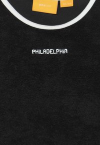 T-shirt de malha preta com decote redondo branco e "PHILADELPHIA" bordado em branco. Etiquetas visíveis em laranja brilhante.