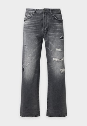 Junger Mann mit rotem T-Shirt mit Logo, abgenutzten grauen Jeans, schwarzen Sneakern, steht mit den Händen in den Taschen vor weißem Hintergrund.