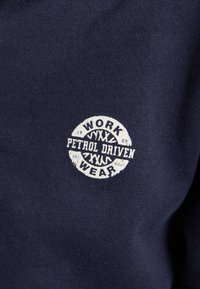 La tela azul marino presenta un logotipo redondo blanco con el texto "WORK WEAR" y "PETROL DRIVEN"; textura suave, diseño casual, detalle centrado.