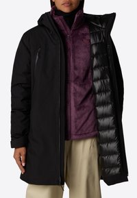 Abrigo exterior negro e impermeable sobre una chaqueta de forro polar morada con cremallera. El abrigo exterior tiene capucha, bolsillos con cremallera y un forro interior acolchado.