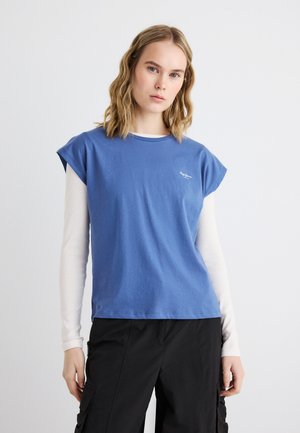 Ung kvinde med bølget blondt hår iført en kortærmet blå T-shirt over en langærmet hvid top og sorte cargobukser, stående mod en hvid baggrund.