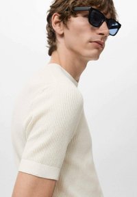 Camisa de punto acanalado beige de manga corta con cuello redondo. El modelo lleva gafas de sol negras. La textura suave resalta el diseño ajustado de la prenda.