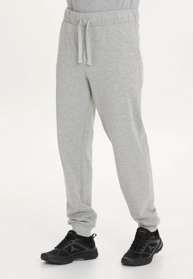 Graue Sweatpants mit elastischem Bund und Kordelzug. Weicher Stoff, schmal zulaufende Beine, Seitentaschen und dezente Branding. Kombiniert mit schwarzen Turnschuhen.