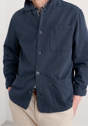 Homme portant une veste bleu marine à boutons avec trois poches avant et manches retroussées, associée à un pantalon beige et une chemise à motifs.