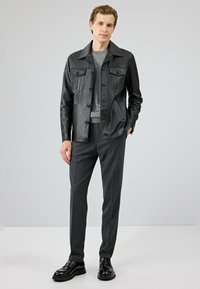 Zwarte leren jas met twee borstzakken, een grijze sweater eronder, donkergrijze op maat gemaakte broek en zwarte glanzende schoenen.