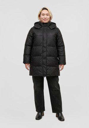 Une femme se tient face à l’objectif, vêtue d’un long manteau noir matelassé, d’un pantalon noir et de bottines, sur un fond uni gris clair.