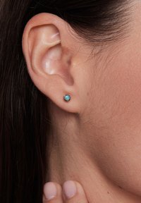 Turkusowe kolczyki na sztyfcie osadzone w złotym metalu. Okrągły kształt z teksturowanym halo wokół centralnego kamienia. Noszone na uchu modelki.