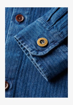 Denimskjorte manchet i en dyb blå nuance med et hæftebreddesle fabric. Har brune knapper og en gylden logo snap detalje. Glat stoffinish.