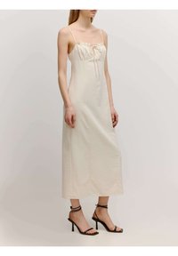 Mittellanges cremefarbenes Kleid mit dünnen Trägern und Kordelzugdetails am Ausschnitt; glatter Stoff und gerade Silhouette. Schwarze Riemchen-Highheels.