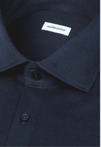 Gros plan d'un col de chemise bleu foncé avec des boutons noirs et une étiquette blanche affichant le nom de la marque "seidensticker."