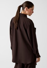 Braunes Langarmhemd mit lockerem Sitz, glatter Textur und geknöpften Manschetten, von hinten gezeigt mit einem seitlichen Pferdeschwanzfrisur.