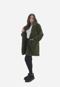 Cappotto in pelliccia ecologica verde con colletto, chiusura a bottoni e tasche laterali, indossato sopra una maglietta bianca, abbinato a leggings neri e stivali.