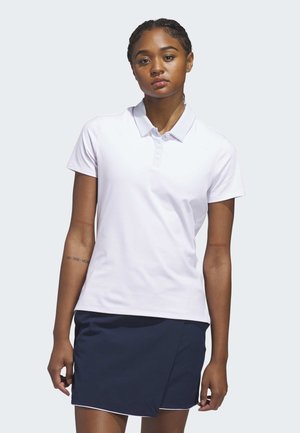 Mladá žena stojící, oblečená v bílé polo košili s krátkými rukávy a tmavě modré sukni na jednoduchém světlém pozadí.