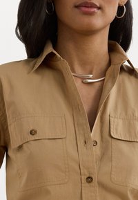 Camicia button-up color tan con due tasche sul petto, in tessuto di cotone, colletto curvo e collana curva argentata indossata attorno al collo.