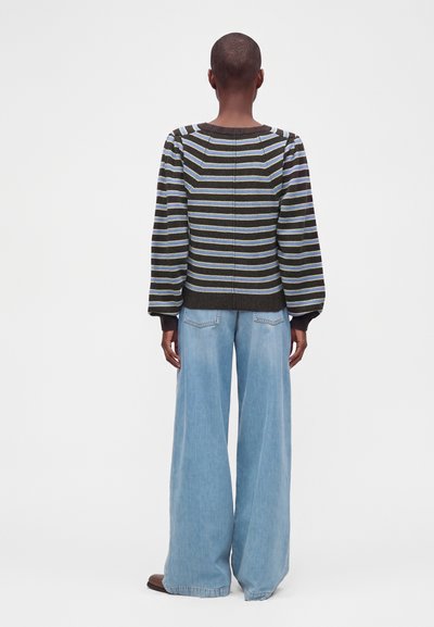 Personne portant un pull rayé et un jean bleu clair à jambes larges, debout de dos sur un fond blanc uni.