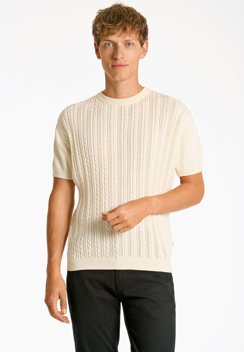 Maglietta a maglia di colore crema chiaro con maniche corte, caratterizzata da un motivo a cavo testurizzato e un collo rotondo a coste. Indossata con pantaloni neri.