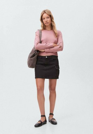 Helles rosa Langarmoberteil, schwarzer Jeansrock mit Seitentasche und schwarze Schuhe. Das Model steht mit verschränkten Armen und einer grauen Tasche über der Schulter.