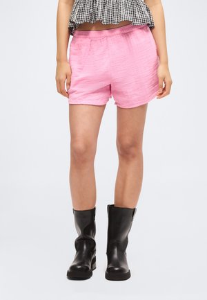 Nő élénk rózsaszín rövidnadrágban, fekete lábszárközépig érő csizmában és fekete-fehér kockás haspólóban, laza karokkal áll.