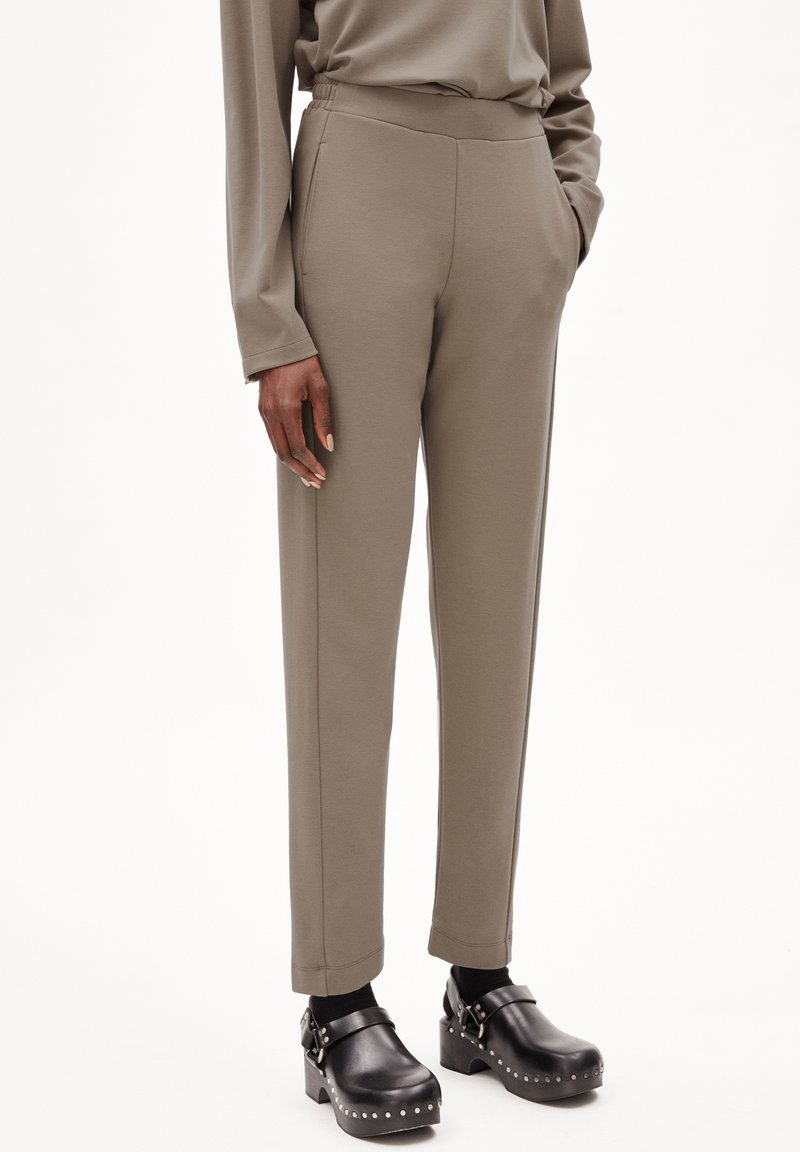 Personne portant un pantalon droit couleur taupe avec poches latérales, un haut assorti, des chaussettes noires et des sabots noirs cloutés sur fond blanc.