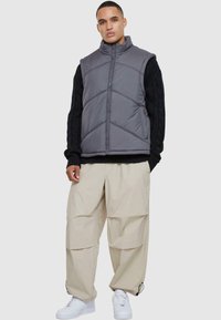 Grå puffväst med hög krage och chevronsöm, bärs över en texturerad svart tröja. Kombineras med beige cargobyxor och vita sneakers.