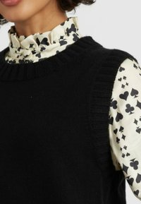 Schwarzer Strickpullover mit gerippten Akzenten, über einer cremefarbenen Bluse mit schwarzem Herzmuster und Rüschenausschnitt getragen.