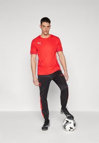 Sarkans sporta krekls ar īsām piedurknēm un Puma logo, savienots ar melniem joggeriem ar sarkaniem sānu svītrām un logo. Spēlētājs stāv uz rakstainas futbola bumbas.