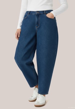 Person iført løse mørkeblå jeans, hvid langærmet top og hvide sneakers, står med den ene hånd i lommen.