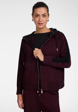 Frau mit dunklem Haar trägt einen bordeauxfarbenen Hoodie mit Reißverschluss, schwarzen Einsätzen und passenden Hosen, posiert vor einem schlichten weißen Hintergrund.