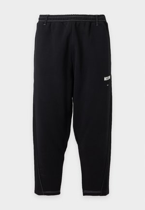 Mand iført hvid Moschino grafisk t-shirt, sorte bukser og sorte sneakers, der står mod en ensartet lys baggrund.
