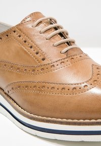 Chaussure brogue en cuir marron clair avec lacets beiges, motifs décoratifs perforés, et semelle blanche avec une bande bleu foncé.