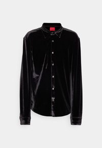 Chemise noire en velours à manches longues avec fermeture boutonnée sur le devant et col pointu, avec une étiquette rouge de la marque HUGO à l’intérieur du col.