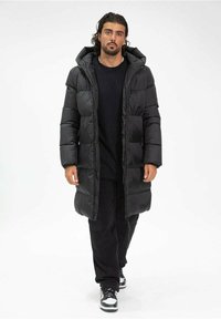 Doudoune noire avec capuche, présentant un design matelassé et une longueur longue. Portée par-dessus une chemise noire et un pantalon noir, avec des baskets.