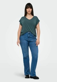 Green v-neck t-shirt with rolled short sleeves, paired with blue flared jeans. Model wears black shoes, standing against a light gray background.