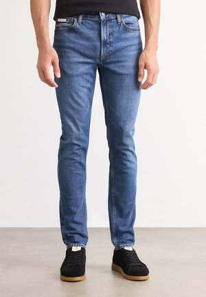 Blauwe spijkerbroeken met een slim fit, voorzien van een klassiek vijf-pocketontwerp en subtiele vervaging. Gecombineerd met zwarte sneakers en een zwart shirt.