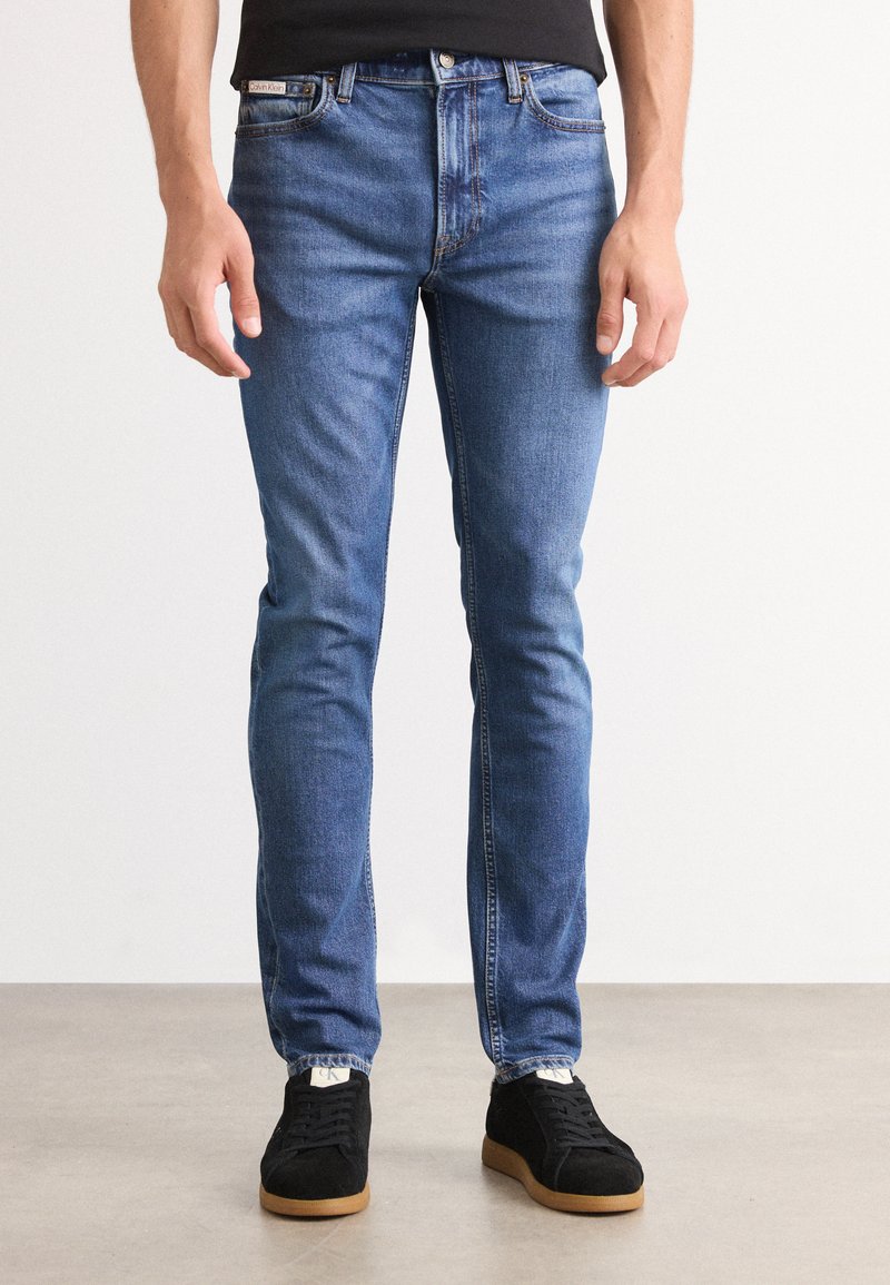 Kék farmernadrág slim fit szabással, klasszikus öt zsebes kialakítással és finom fakulással. Fekete sportcipővel és fekete pólóval párosítva.