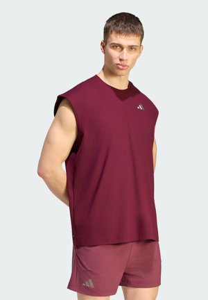 Junger Mann, der ein ärmelloses bordeauxfarbenes Sportshirt und passende Shorts trägt, steht vor einem einfarbigen hellen Hintergrund.