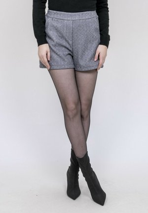 Personne portant un short gris taille haute, des collants noirs transparents et des bottes noires à talons aiguilles pointus, debout jambes croisées.