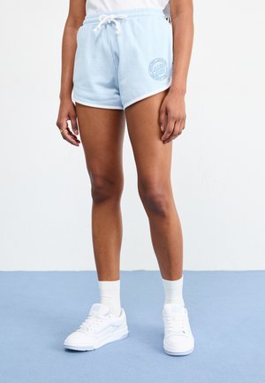 Shorts en coton bleu clair avec des bordures blanches ; la cuisse gauche présente un logo circulaire "Santa Cruz". Associé à des baskets blanches et des chaussettes mi-hautes.