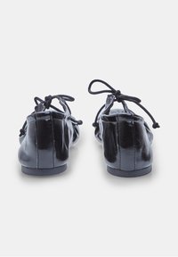Musta nahka ballet-tossu, jossa on kiiltävä pinta. Ominaisuuksiin kuuluu pyöristetty kärki, pieni korko ja säädettävät nauhat takana.