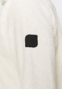 Weißer Fleecejacke mit einer weichen Textur und einem schwarzen Lederlogo-Patch auf der linken Seite. Das Design ist einfach und gemütlich.