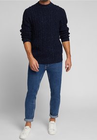 Granatowy sweter w splot kablowy z wielokolorowymi wstawkami, okrągły dekolt, noszony z niebieskimi dżinsami z podwiniętymi mankietami i białymi trampkami.