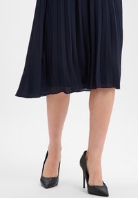 Granatowa plisowana spódnica midi o zwiewnym kroju, zestawiona z czarnymi szpilkami z noskiem w szpic i gładką fakturą skóry.