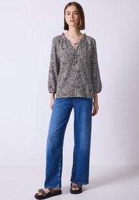Jeune femme portant un chemisier floral à manches longues, un jean large bleu et des sandales noires à boucles, debout devant un fond uni.