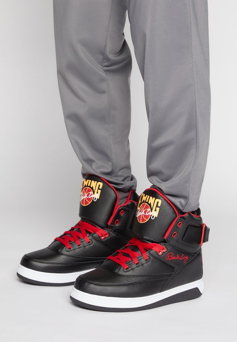 Des baskets montantes noires avec des lacets et des accents rouges, une texture en cuir et un col rembourré. Logo sur la languette et le côté.