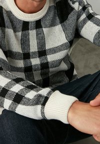 Suéter de cuadros en blanco y negro con un cuello y puños de crema acanalados, que presenta un tejido texturizado. Las manos del modelo descansan sobre las rodillas.