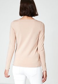 Kobieta z brązowymi włosami sięgającymi ramion, ubrana w beżową bluzkę z długim rękawem i białe spodnie, przedstawiona z tyłu na jednolitym tle.