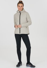 Vaalea harmaa eristetty takki huppulla ja etuvetoketjulla, yhdistetty mustiin legginsseihin ja urheilukenkihin, valkoista taustaa vasten.
